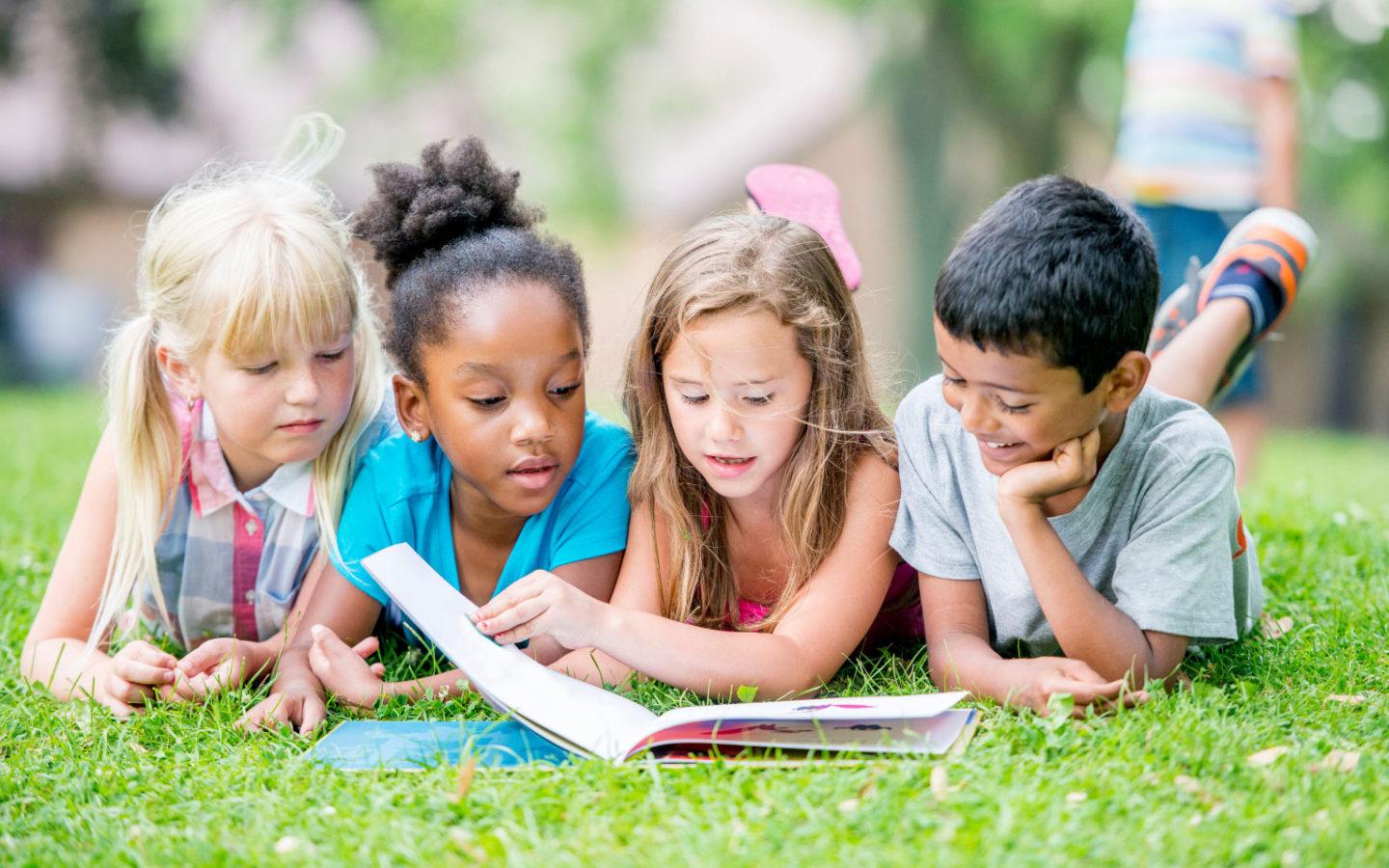 four children lying on grass and reading a book together